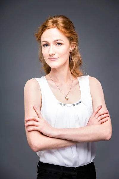 Retrato de una joven pelirroja hermosa chica en el estudio sobre un fondo gris aislado. Una mujer está de pie con los brazos cruzados y sonriendo con una camisa blanca con una manga corta. Concepto empresarial — Foto de Stock