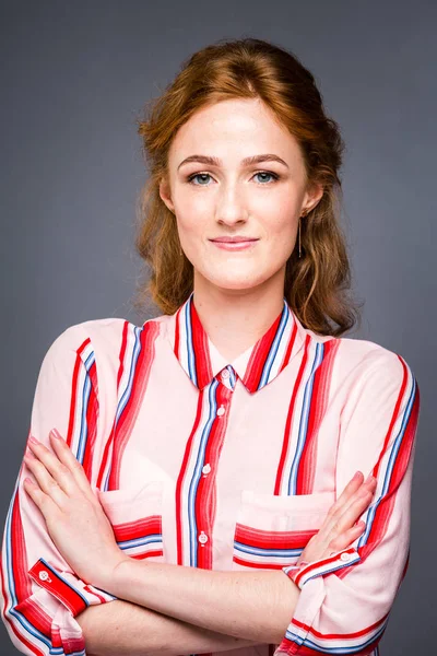 Retrato de una joven chica hermosa pelirroja en el estudio sobre un fondo gris aislado. Una mujer se para y sonríe con una camisa roja con una manga corta. Concepto empresarial — Foto de Stock