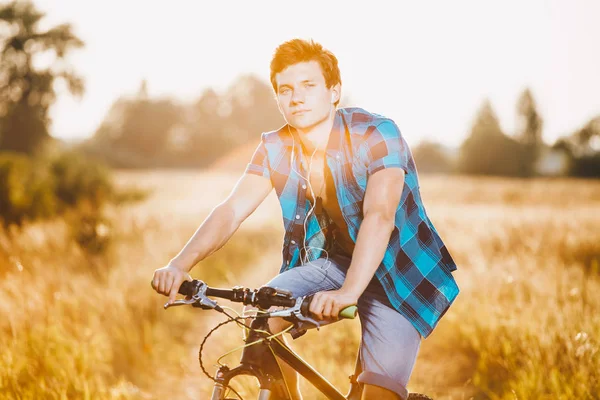 A young guy in a shirt and naked torso sits on a bicycle in headphones and listens to music outdoors, nature, field outside city — Stock Photo, Image