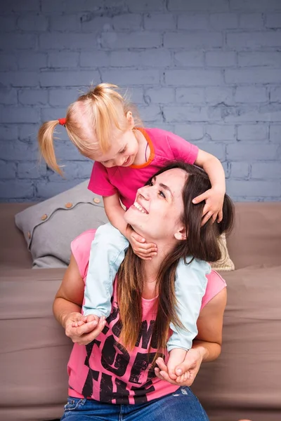 Uma jovem e esbelta mãe bonita com sua filha loira em seus ombros jogar, desfrutar e entrar nas equipes perto do sofá no chão de madeira — Fotografia de Stock