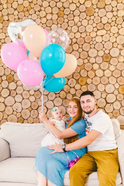 Junger Familienvater, Frau und kleines Mädchen sitzen auf der Couch im Haus mit dekorativer Wand aus Holz und bunten Luftballons mit Helium in der Hand. — Stockfoto