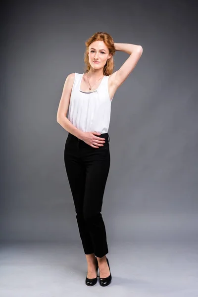 Bela jovem mulher caucasiana com longos cabelos vermelhos em saltos altos, calças pretas e uma camisa branca está em pleno crescimento em um fundo isolado cinza . — Fotografia de Stock