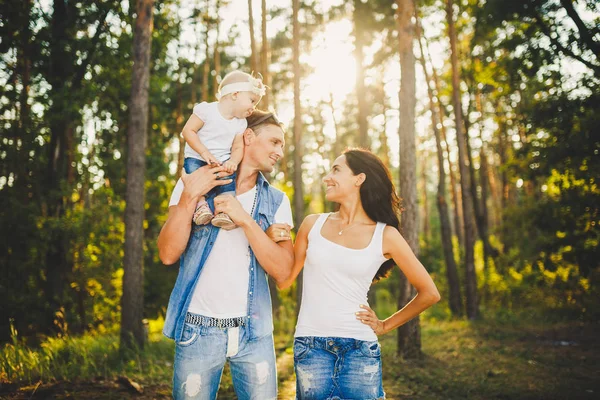 Stock image stylish young Family of mom, dad and daughter one year old blonde sitting near father on shoulders, outdoors outside the city in a park amid tall trees in summer at sunset.