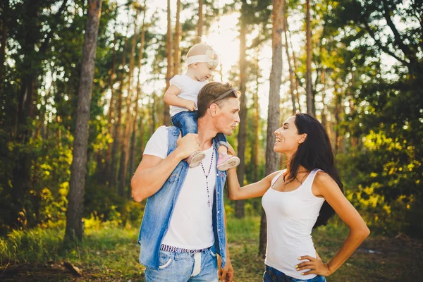 Snygg ung familj av mamma, Pappa och dotter ett år gammal blond sitter nära far på axlar, Utomhus utanför staden i en park mitt i höga träd på sommaren vid solnedgången. — Stockfoto