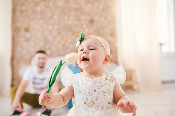 Lilla baby flicka blondin med ett bandage på huvudet håller en färg tulbpin inne i huset på bakgrund av hennes föräldrar. — Stockfoto