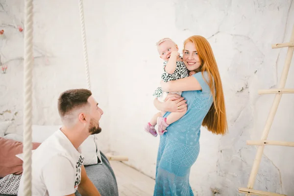 Jeune belle femme aux cheveux longs rouges dans une robe bleue tient un enfant sur ses mains un an blonde près du lit sur lequel repose un homme reposant — Photo