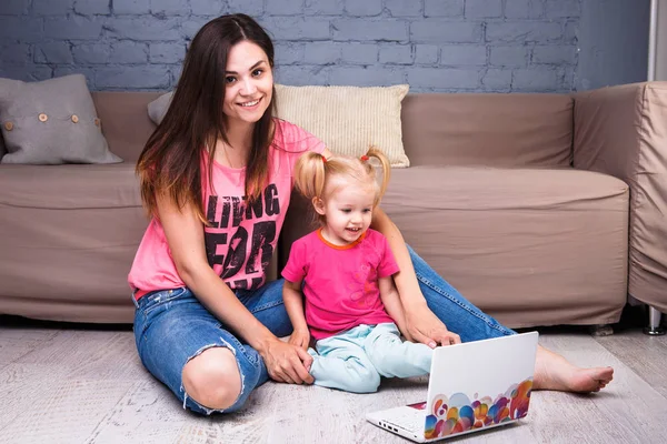 Güzel genç şık beyaz anne ile sarışın kızı odanın içinde odasında bir ahşap zemin kanepede oturan bir taşınabilir bilgisayar kullanarak — Stok fotoğraf