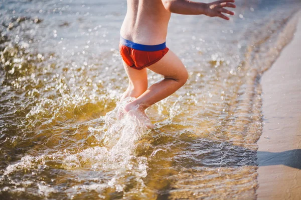 Wesoły dziecko płci męskiej trzech lat biegnącej wzdłuż plaży w pobliżu wody i rozpryskiwania latać. Aktywny wypoczynek w lato w pobliżu rzeki — Zdjęcie stockowe
