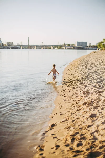 Un niño alegre de tres años corriendo por la playa cerca del agua y salpicando la mosca. Recreación activa En verano cerca del río —  Fotos de Stock