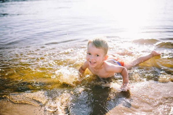Ein kaukasisches Kind von drei Jahren liegt in roter Badehose auf dem Bauch im Wasser am Flussufer eines Sandstrandes. lernt mit einem Lächeln schwimmen — Stockfoto