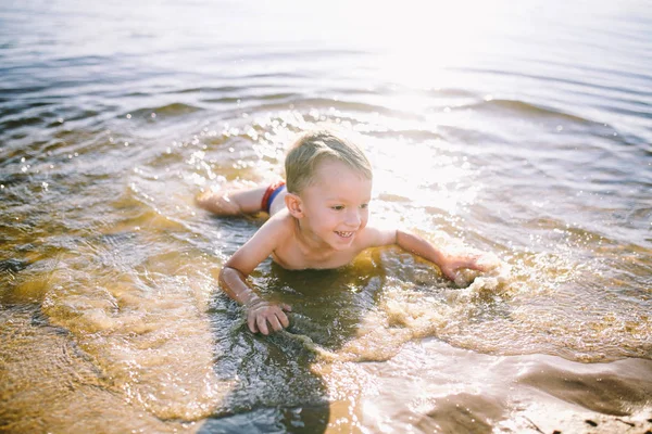 Ein kaukasisches Kind von drei Jahren liegt in roter Badehose auf dem Bauch im Wasser am Flussufer eines Sandstrandes. lernt mit einem Lächeln schwimmen — Stockfoto