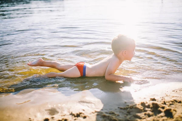 Un enfant caucasien de trois ans dans des troncs de natation rouges se trouve sur le ventre dans l'eau près de la rive d'une plage de sable fin. Apprend à nager avec le sourire — Photo