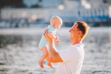 Genç bir baba oğlunun bir yıl bir gülümseme ile onun kollarında tutar ve hugs seviyor ve onu sahilde nehir kıyısında atar
