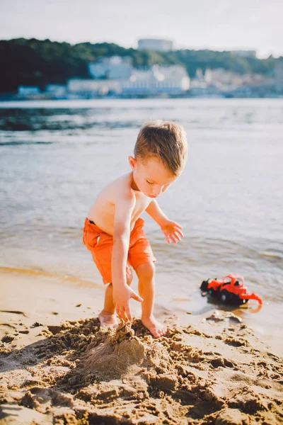 Un petit garçon construit des figures de sable sur la rive de la rivière au coucher du soleil le jour, les mains font des formes à partir de sable humide — Photo