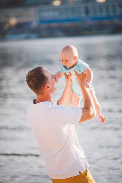 Een jonge vader houdt van één jaar van zijn zoon in zijn armen met een glimlach en houdt van knuffels en gooit hem op de rivieroever op het strand — Stockfoto