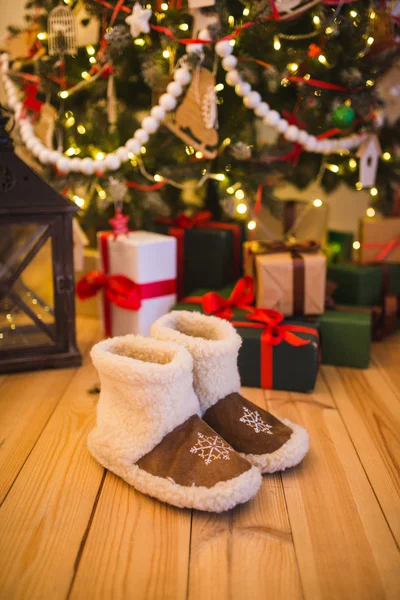 Ζευγάρι ζεστό σπίτι παπούτσια κατασκευασμένα από μαλλί που στέκεται κοντά το χριστουγεννιάτικο δέντρο με δώρα στο σπίτι στο ξύλινο πάτωμα — Φωτογραφία Αρχείου