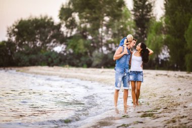 Üç kişi anne, baba ve kızı deniz kıyısı yalınayak elini tutarak yürüyen bir adam omuzlarında bir yılın genç güzel ve şık giyimli ailesi