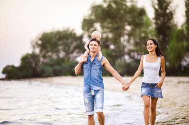 Üç kişi anne, baba ve kızı deniz kıyısı yalınayak elini tutarak yürüyen bir adam omuzlarında bir yılın genç güzel ve şık giyimli ailesi