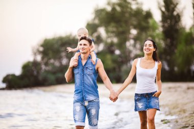 Üç kişi anne, baba ve kızı deniz kıyısı yalınayak elini tutarak yürüyen bir adam omuzlarında bir yılın genç güzel ve şık giyimli ailesi