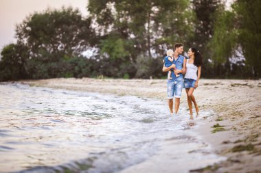 Üç kişi anne, baba ve kızı deniz kıyısı yalınayak elini tutarak yürüyen bir adam omuzlarında bir yılın genç güzel ve şık giyimli ailesi
