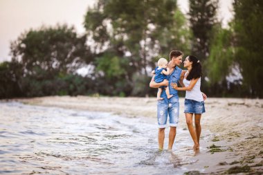 Üç kişi anne, baba ve kızı deniz kıyısı yalınayak elini tutarak yürüyen bir adam omuzlarında bir yılın genç güzel ve şık giyimli ailesi