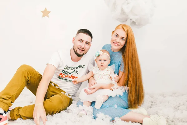 A group of young people, a family of father and mother 30 years and a daughter of one year, houses in a light interior sit on a wooden floor, a background of a white wall and artificial snow. — Stock Photo, Image