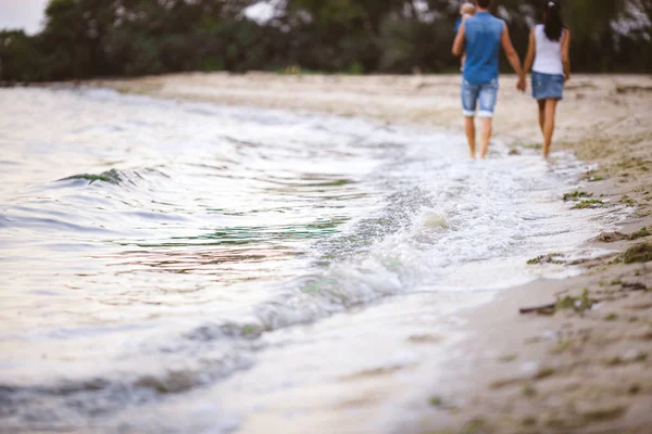 Vacaciones familiares cerca del mar. Dos adultos en ropa vaquera con un niño en sus brazos caminan por la playa de arena por detrás —  Fotos de Stock