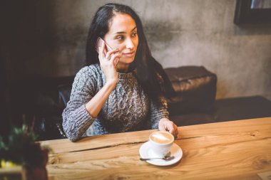 ahşap masa pencere yakınındaki cep telefonu üzerinde konuşurken ve kafede kahve içme güzel genç esmer kız Noel dekor ile dekore edilmiştir. Bir gri örgü yün kazak giymiş