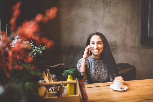 Hermosa chica morena joven hablando en el teléfono móvil en la mesa de madera cerca de la ventana y beber café en la cafetería decorada con decoración de Navidad. Vestido con un suéter de lana de punto gris — Foto de Stock