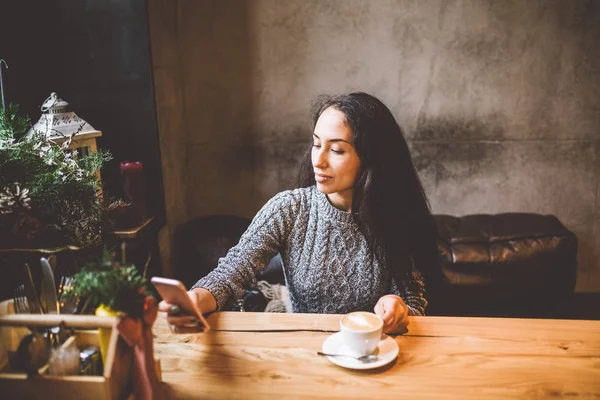 Bella ragazza usa, scrive testo su un telefono cellulare a un tavolo di legno vicino alla finestra e beve caffè in un caffè decorato con decorazioni natalizie. Vestita con un maglione grigio di lana lavorato a maglia — Foto Stock