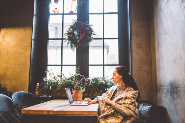Hermosa niña utiliza la tecnología portátil, tipos de texto mirando el monitor en un café junto a la ventana en la mesa de madera, en invierno decorado con decoración de Navidad.Vestido con un suéter de lana de punto gris — Foto de Stock
