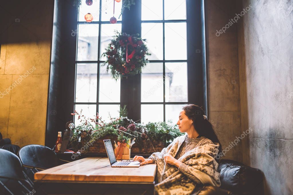 beautiful young girl uses laptop technology, types text looking at the monitor in a cafe by the window at wooden table, in winter decorated with Christmas decor.Dressed in a gray knitted wool sweater