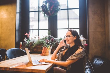 Güzel genç esmer kadının içinde café Noel iç ahşap masa pencerenin telefonda konuşurken ve laptop arıyorum. Kışın, o bir gri örgü kazak ve gözlük giymiş