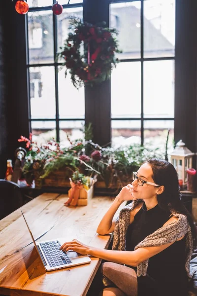 Όμορφη νεαρή μελαχρινή γυναίκα μέσα στο καφενείο με εσωτερικό Χριστούγεννα στο ξύλινο τραπέζι δίπλα στο παράθυρο μιλώντας στο τηλέφωνο και να ψάχνει το lap-top. Το χειμώνα, αυτή είναι ντυμένος με ένα γκρι πλεκτό πουλόβερ και γυαλιά — Φωτογραφία Αρχείου