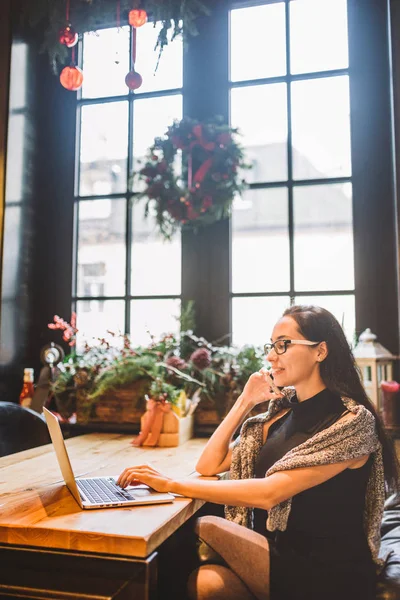 Mulher morena bonita dentro do café com interior de Natal na mesa de madeira pela janela falando no telefone e olhando o laptop.In inverno, ela está vestida com uma camisola de malha cinza e óculos — Fotografia de Stock