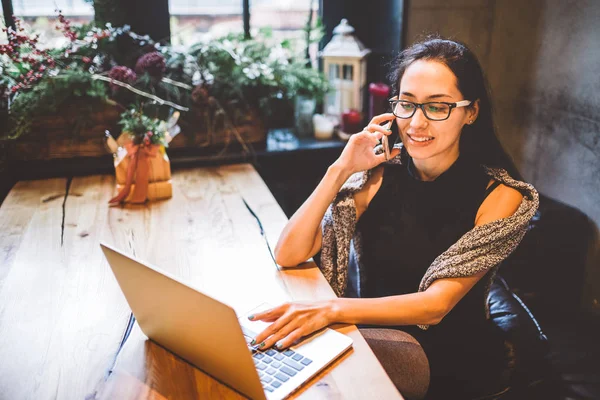 Mooie jonge brunette vrouw binnen café met Kerstmis interieur aan houten tafel bij het raam praten over telefoon en op zoek naar de laptop. In de winter, is ze gekleed in een grijze gebreide trui en glazen — Stockfoto