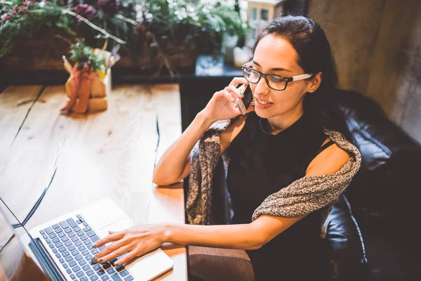 Mooie jonge brunette vrouw binnen café met Kerstmis interieur aan houten tafel bij het raam praten over telefoon en op zoek naar de laptop. In de winter, is ze gekleed in een grijze gebreide trui en glazen — Stockfoto