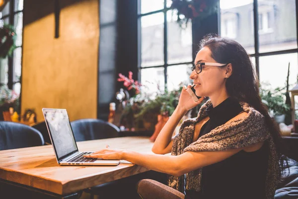 Mooie jonge brunette vrouw binnen café met Kerstmis interieur aan houten tafel bij het raam praten over telefoon en op zoek naar de laptop. In de winter, is ze gekleed in een grijze gebreide trui en glazen — Stockfoto