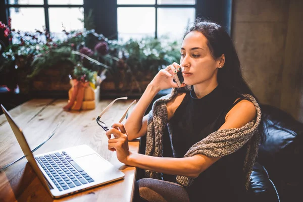 Mulher morena bonita dentro do café com interior de Natal na mesa de madeira pela janela falando no telefone e olhando o laptop.In inverno, ela está vestida com uma camisola de malha cinza e óculos — Fotografia de Stock