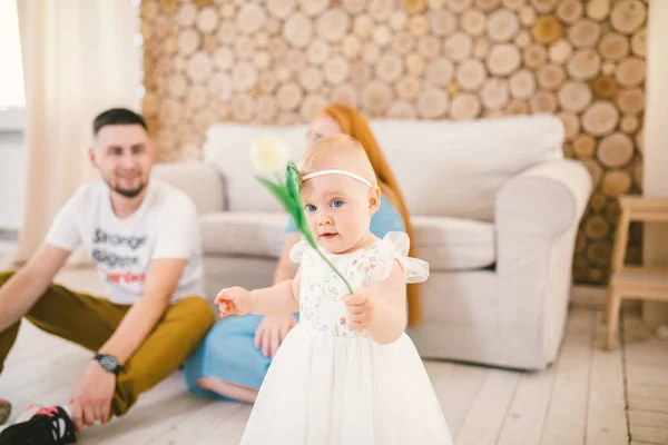 Jong gezin zit op de grond in de buurt van de Bank, kleine dochter van een één-jarige blondine is te leren om te lopen in een witte jurk tegen de achtergrond van de gelukkige ouders. Het concept van de familie vakantie huis — Stockfoto