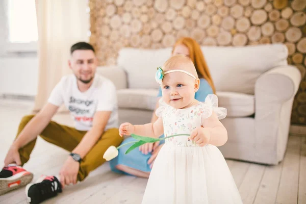 Junge Familie sitzt auf dem Boden in der Nähe der Couch, kleine Tochter einer blonden Einjährigen lernt im weißen Kleid vor dem Hintergrund glücklicher Eltern zu gehen. — Stockfoto