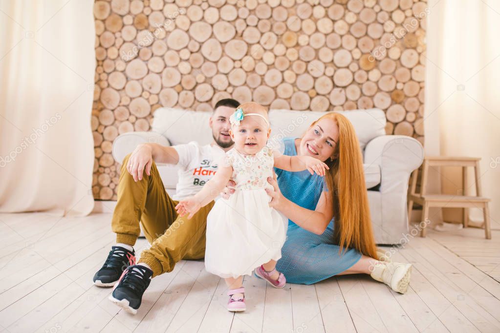 young family sits on the floor near the couch,small daughter of a blonde one year old is learning to walk in a white dress against the background of happy parents.The concept of family vacation home