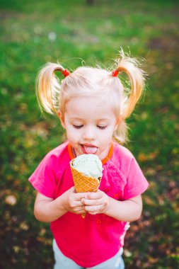 little funny Caucasian girl blonde with blue eyes with two tails on head eating an ice cream in a waffle cup of blue sitting on green grass. All face dirty in melted ice cream.Looks down at the camera clipart