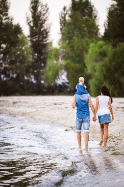 Aile tatil deniz kenarında. Arkadan kumlu plaj boyunca onların kollarında bir çocukla kot pantolon giysili iki yetişkin yürümek