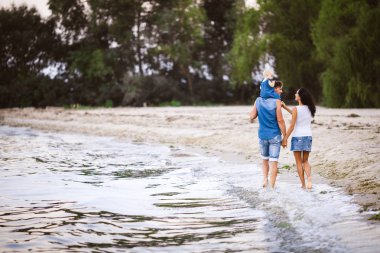 Aile tatil deniz kenarında. Arkadan kumlu plaj boyunca onların kollarında bir çocukla kot pantolon giysili iki yetişkin yürümek