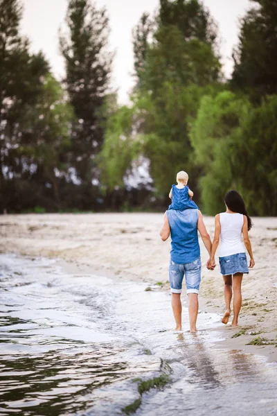 Vacaciones familiares cerca del mar. Dos adultos en ropa vaquera con un niño en sus brazos caminan por la playa de arena por detrás —  Fotos de Stock