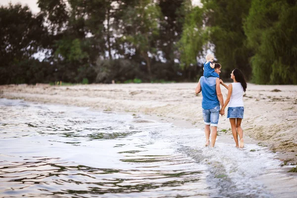 Vakantie met het gezin in de buurt van de zee. Twee volwassenen in jeans kleding met een kind in hun armen lopen langs het zandstrand van achter — Stockfoto