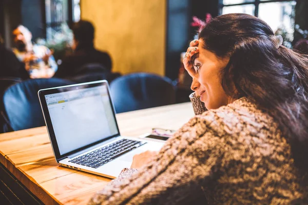 Mooie jonge brunette meisje in trui werken op laptopcomputer binnen café aan houten tafel in de buurt van venster. Winter en Kerstmis decoratie. — Stockfoto