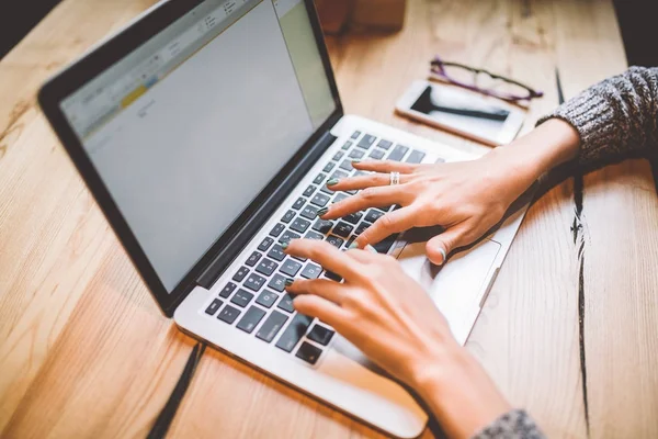 El primer plano de la mano de una joven en un suéter gris utiliza la tecnología de computadora portátil en una mesa de madera en un café. Cerca del teléfono móvil y gafas. El concepto de trabajo remoto dama de negocios . — Foto de Stock