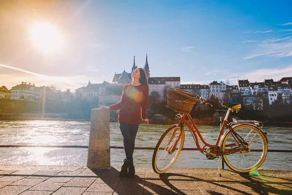 Uma bela jovem está no aterro perto de uma bicicleta da cidade com uma cesta de vermelho na Suíça, a cidade de Basileia. Dia ensolarado na velha Europa — Fotografia de Stock